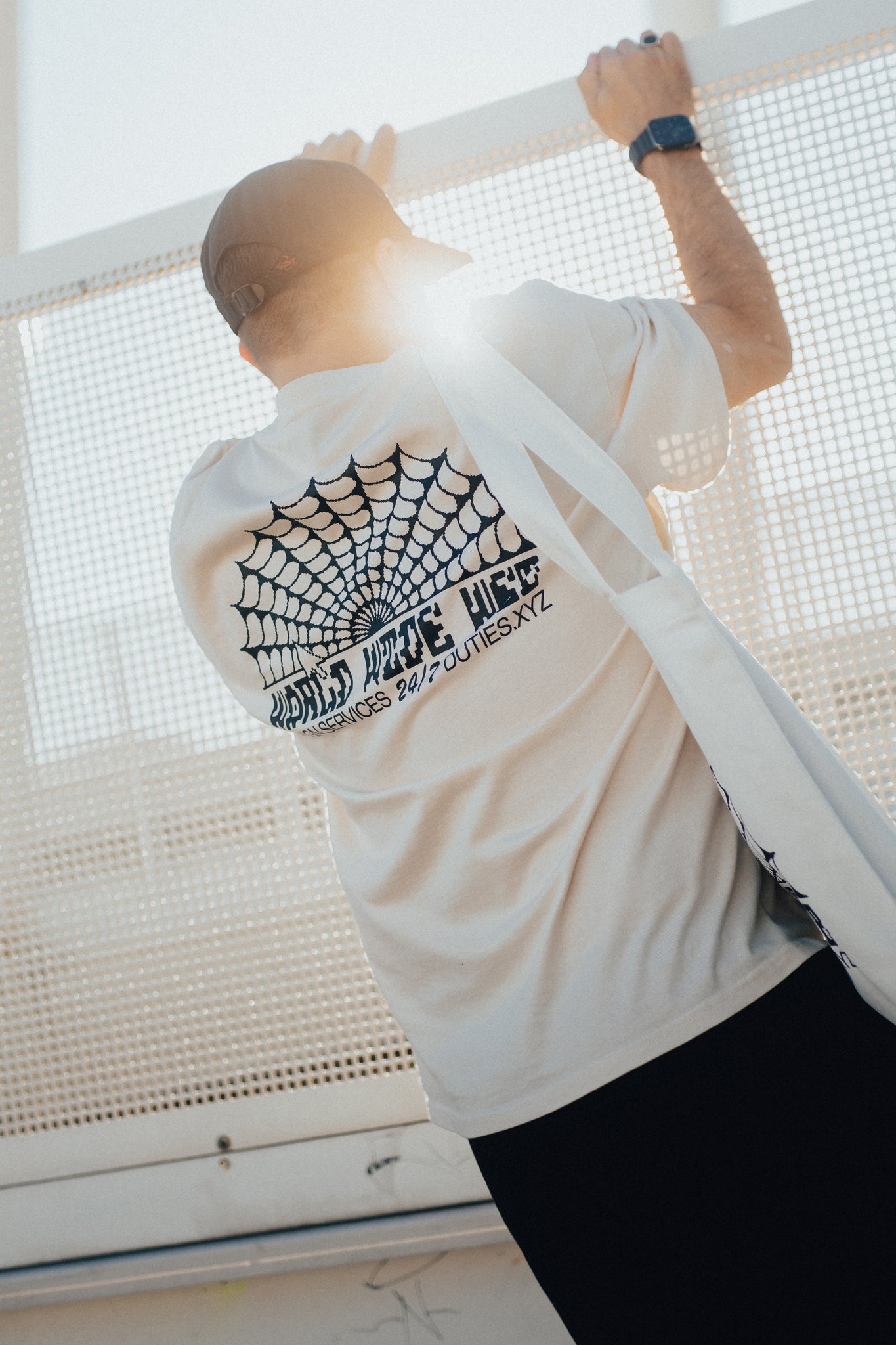 Man wearing an Ecru-colored World Wide Web T-shirt while climbing on a fence with his back against the camera showcasing the black printed graphics.