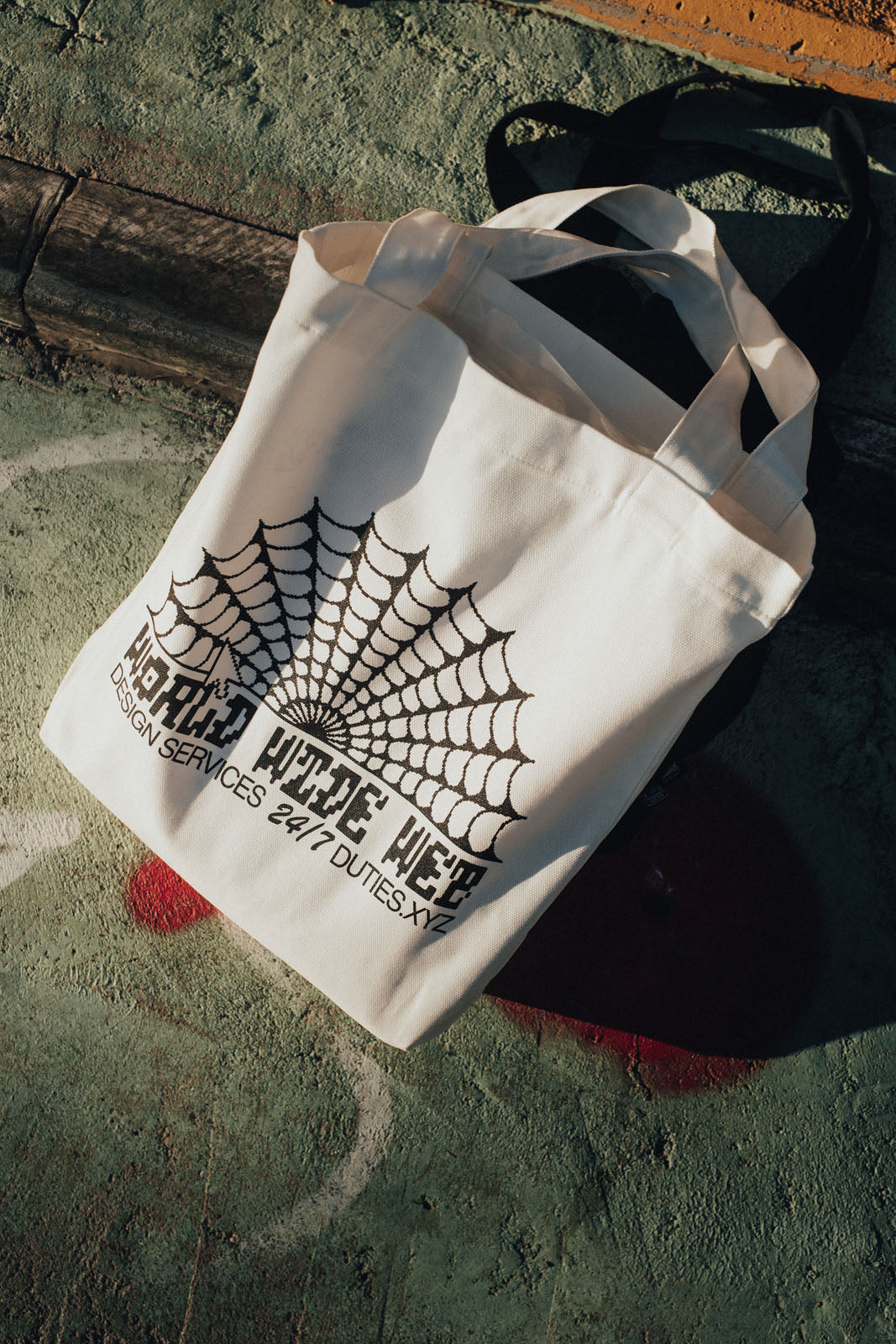 Photo of the Ecru-colored Duties World Wide Web tote bag from above, showcasing the black graphic print. The tote bag is standing on a green colored concrete surface.