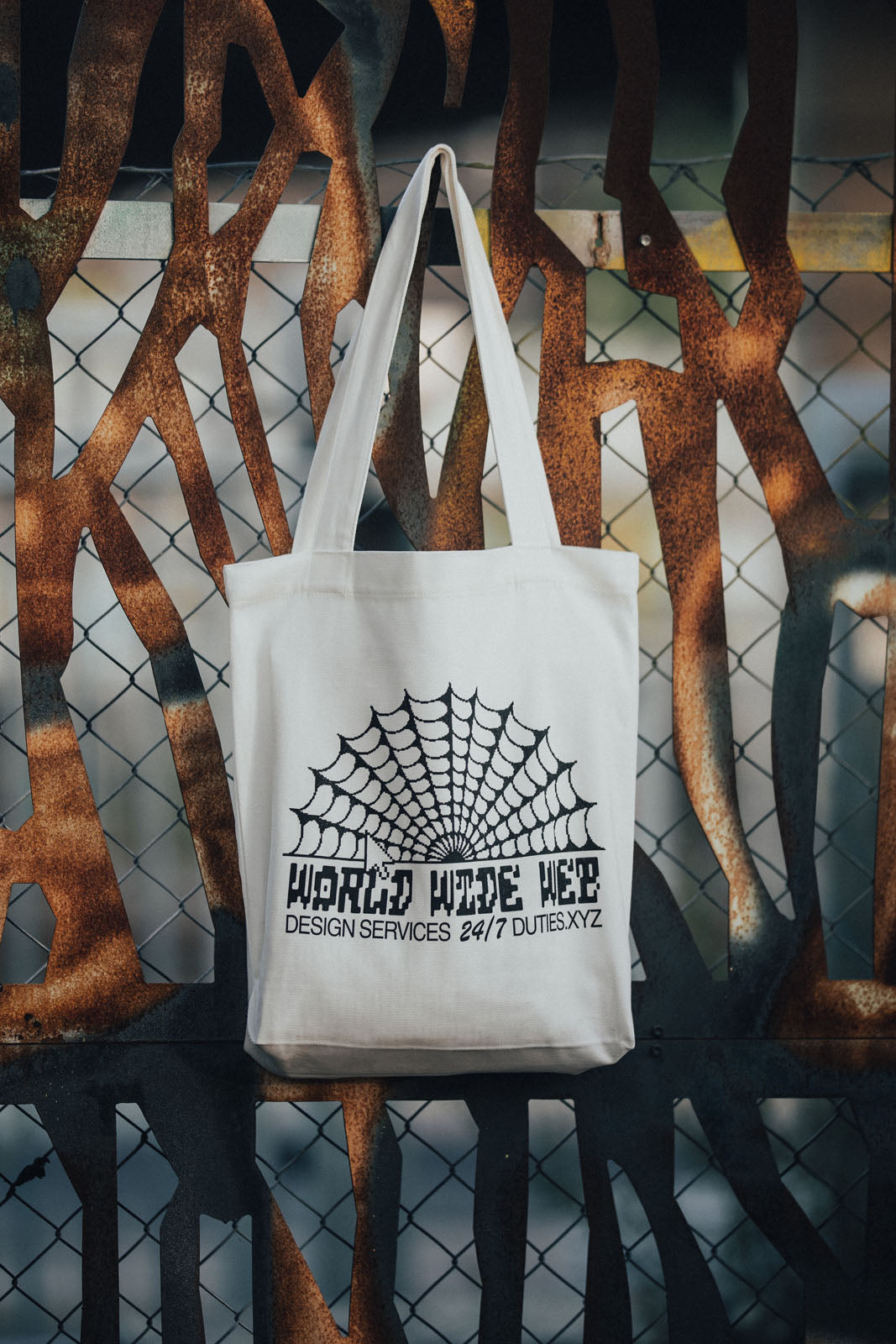 Photo of the Ecru-colored World Wide Web tote bag hanging on a rusty fence, showcasing the black printed Duties World Wide Web graphic on the fabric.