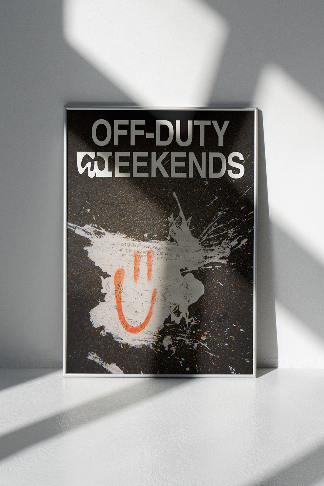 Framed Duties Smiley print poster featuring a spray-painted smiley face on a white paint splash on the ground, with text saying 'OFF-DUTY WEEKENDS', standing on a white floor and leaning against a white wall.